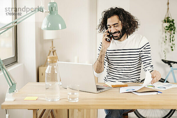 Fröhlicher junger Mann telefoniert  während er von zu Hause aus arbeitet