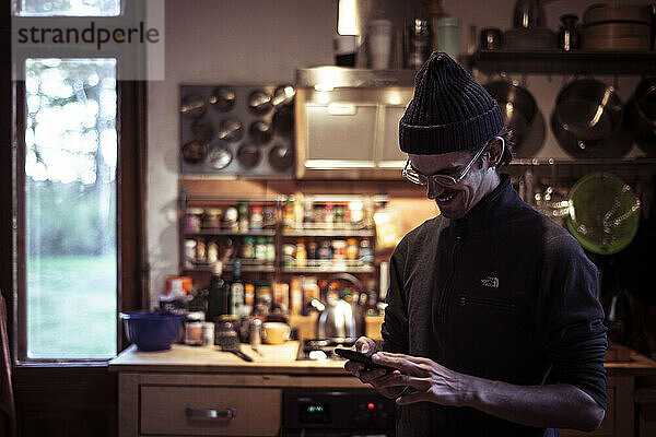 Junger lächelnder Mann mit Brille und Mütze überprüft sein Mobiltelefon in der Küche