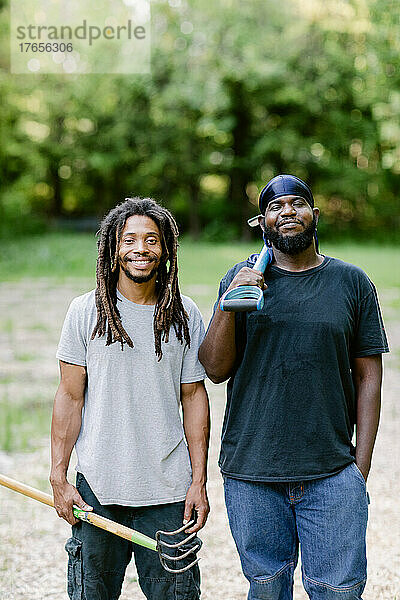 Zwei schwarze Bauern lächeln in die Kamera  während sie landwirtschaftliche Werkzeuge in der Hand halten