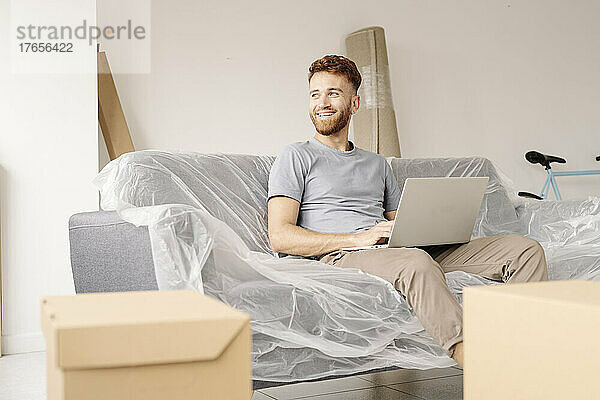 Ein junger Mann sitzt auf seiner in Plastik eingewickelten Couch und benutzt seinen Laptop.
