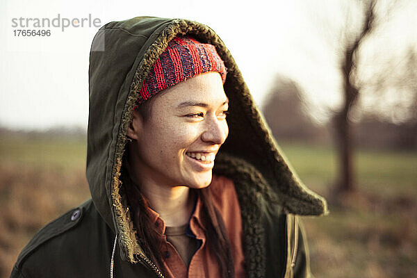 Nicht-binäre Person in warmer Jackenkapuze lächelt draußen in der Nachmittagssonne