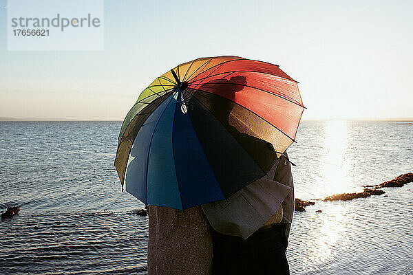 Lesbisches Paar küsst sich unter einem Pride-Regenschirm am Strand