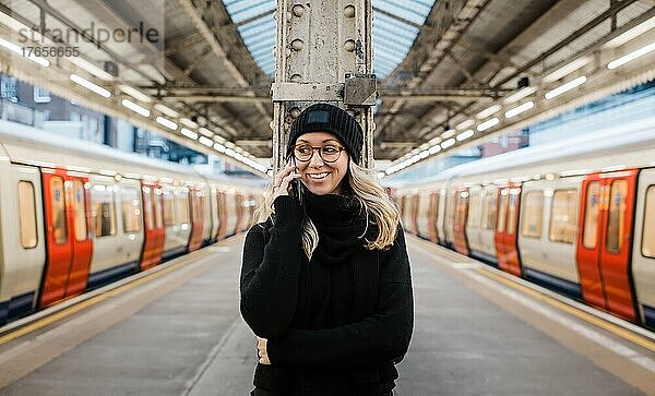 Frau auf einem Bahnsteig  die auf ihrem Telefon spricht und auf einen Londoner Zug wartet