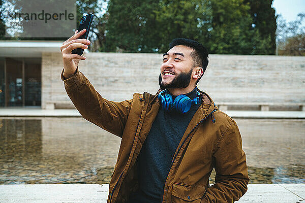 Fröhlicher asiatischer Mann in Oberbekleidung  der ein Selfie mit dem Smartphone macht