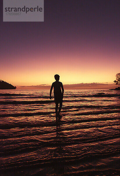 Silhouette eines Jungen  der bei Sonnenuntergang im seichten Wasser am Strand steht.