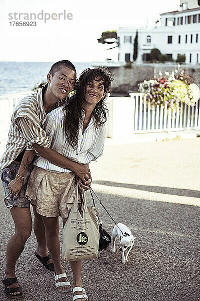 Ein queeres weibliches Paar umarmt sich lachend mit Hunden am Meer
