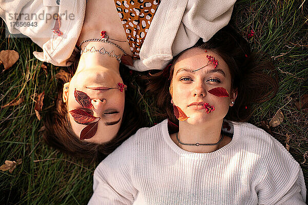 Zwei junge Frauen mit roten Blättern im Gesicht liegen im Herbst im Gras