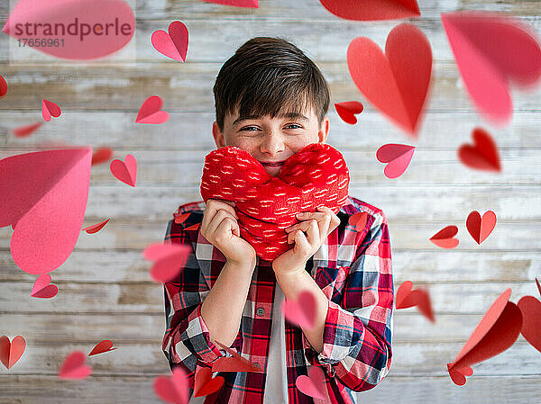 Netter Junge hält ein Herzkissen  umgeben von schwebenden Papierherzen.