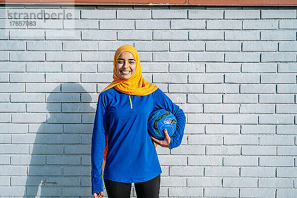 Fröhliche islamische Frau mit Ball in der Nähe einer Ziegelmauer