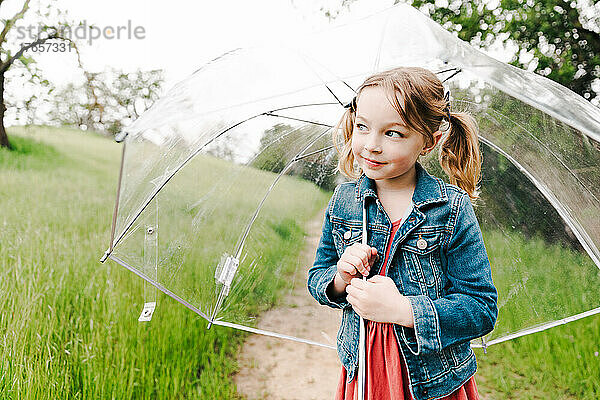 Kleines Mädchen mit Zöpfen  das im Frühling einen durchsichtigen Regenschirm im Freien hält