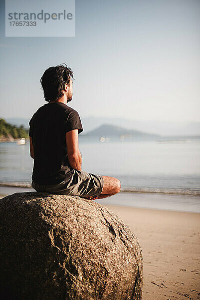 Mann sitzt auf einem Felsen und meditiert am Strand