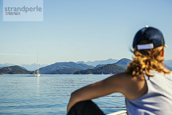 Eine Frau blickt auf eine ruhige Seeszene mit Segelboot und Bergen