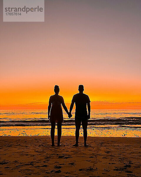 Silhouette eines Paares  das Händchen hält und den Sonnenuntergang am Strand beobachtet.