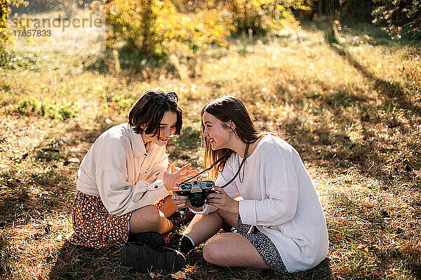 Fröhliche Freundinnen mit Kamera sitzen im Herbst auf Gras im Park
