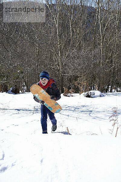 Fröhlicher Teenager läuft auf Schnee und hält ein Surfbrett in der Hand