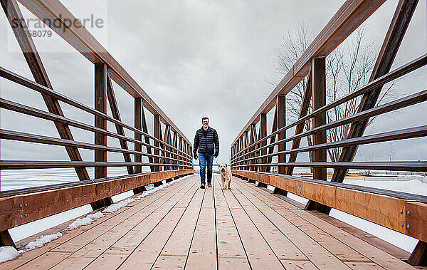Mann geht an bewölktem Wintertag mit flauschigem Hund über eine Brücke.