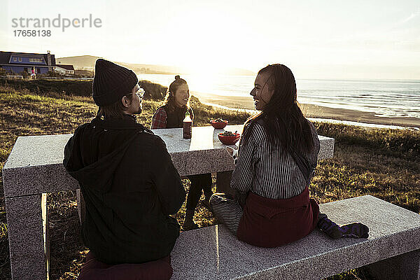 Freunde sitzen bei Sonnenuntergang am Meer und reden und essen