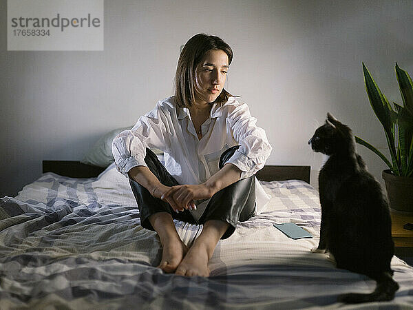 Frau sitzt abends mit einer Katze auf dem Bett