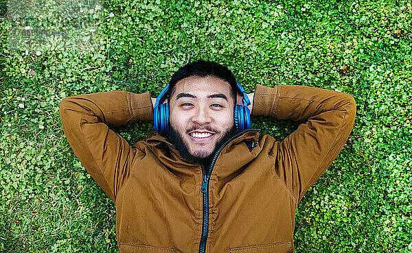 Glücklicher ethnischer Mann  der Musik mit Kopfhörern auf grünem Gras genießt