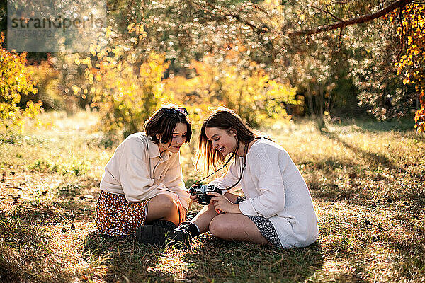 Zwei Mädchen im Teenageralter sitzen im Herbst mit Kamera auf Gras im Park