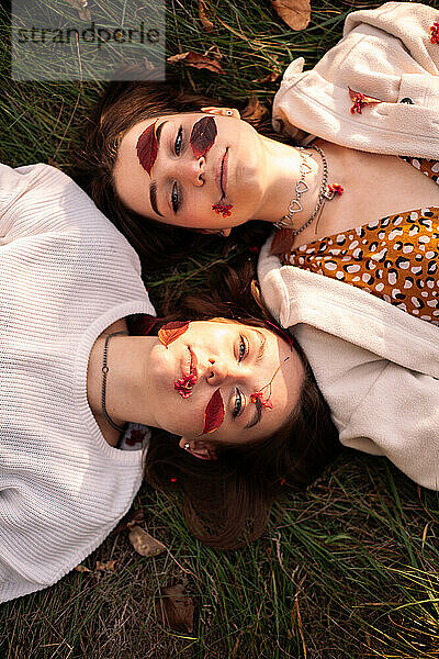 Zwei Mädchen im Teenageralter mit roten Blättern im Gesicht liegen im Herbst im Gras