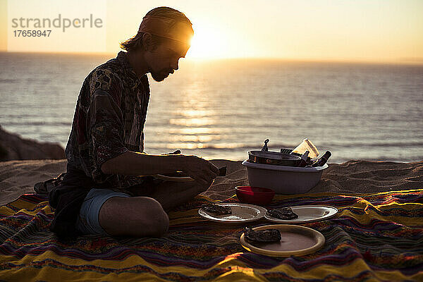 Sonnenuntergang über dem Meer  während ein Mann im Sand Portugals ein Picknick macht