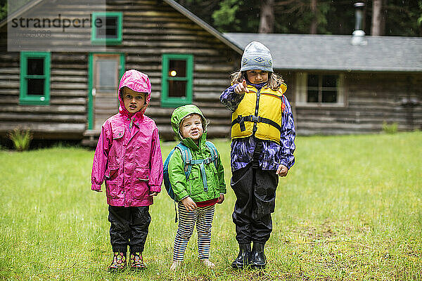 Drei kleine Kinder in Regenkleidung vor einer Blockhütte