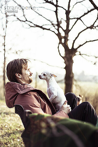 Glücklicher junger Mann mit Jacke sitzt draußen mit Hund auf dem Schoß in der Sonne