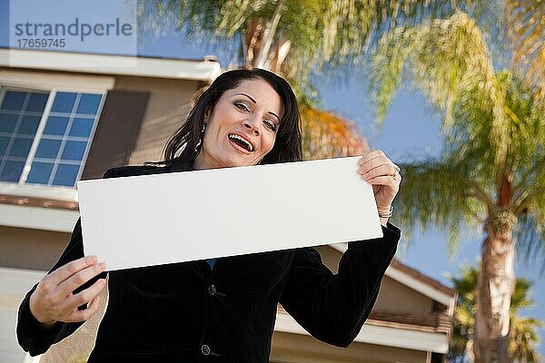 Glückliche attraktive hispanische Frau hält leeres Schild vor einem Haus