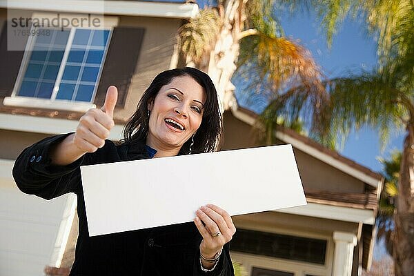 Glückliche attraktive hispanische Frau mit Daumen hoch  die ein leeres Schild vor einem Haus hält