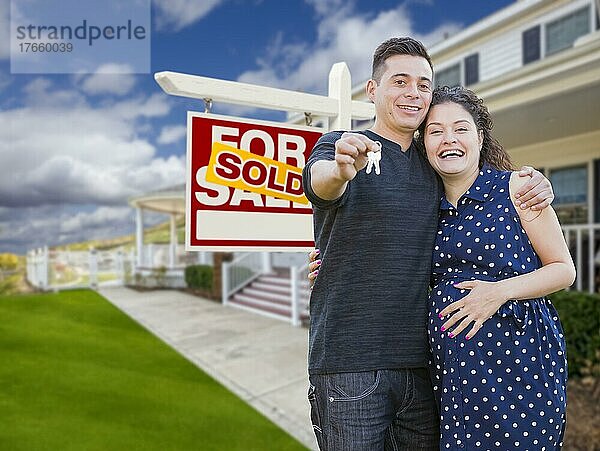 Glückliches hispanisches Paar vor neuem Haus und verkauftem Immobilienschild  das seine Hausschlüssel zeigt