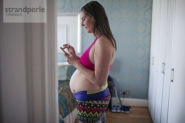 Schwangere Frau im Sport-BH benutzt ihr Smartphone in der Haustür