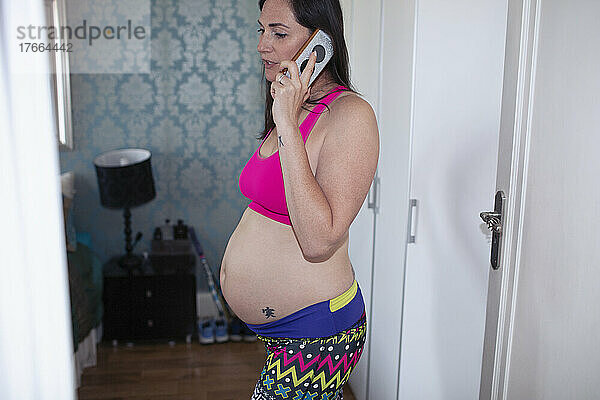 Schwangere Frau im Sport-BH  die zu Hause mit ihrem Smartphone telefoniert