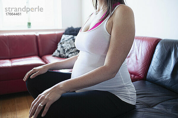 Schwangere Frau im Tanktop meditiert auf dem Wohnzimmersofa