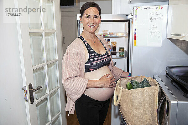 Portrait lächelnde schwangere Frau im Sport-BH beim Ausladen von Lebensmitteln