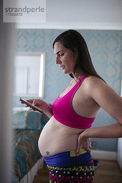 Schwangere Frau im Sport-BH benutzt ihr Smartphone in der Tür