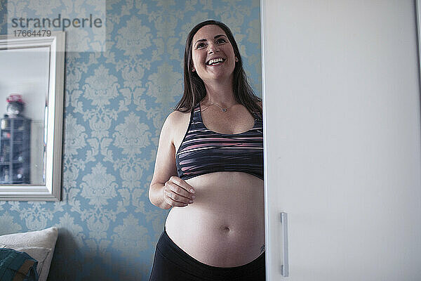 Glückliche schwangere Frau im Sport-BH am Schlafzimmerschrank