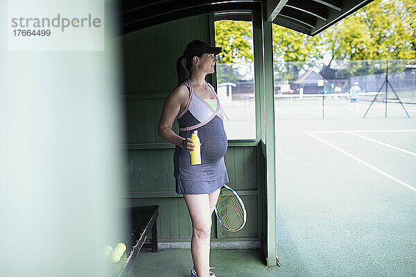 Schwangere Frau im Kleid trinkt Wasser auf dem Tennisplatz