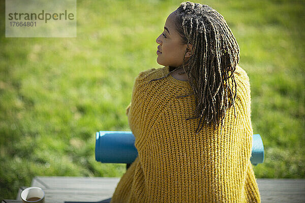 Junge Frau im Pullover mit Yogamatte auf sonniger Terrasse