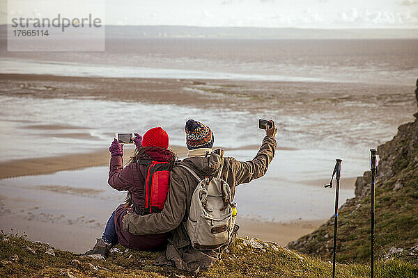 Wandererpaar mit Fotohandys auf einer Klippe mit Blick auf den Winterstrand