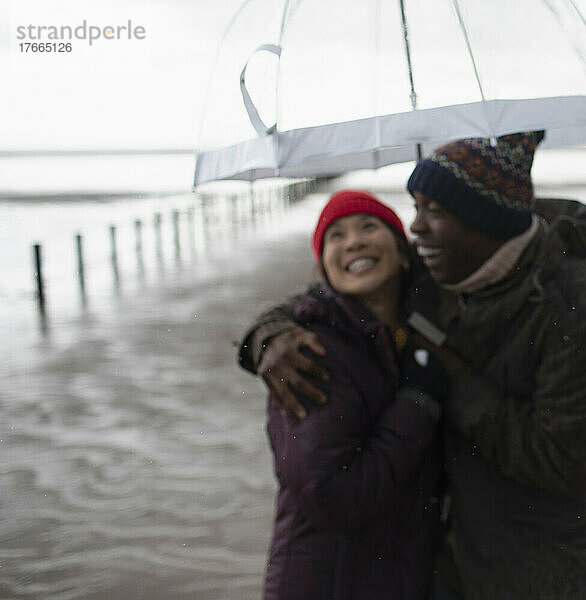 Glückliches Paar in warmer Kleidung unter Sonnenschirm am nassen Winterstrand