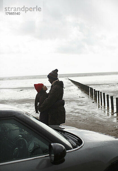 Zärtliches Paar in warmer Kleidung  das sich am nassen Winterstrand umarmt