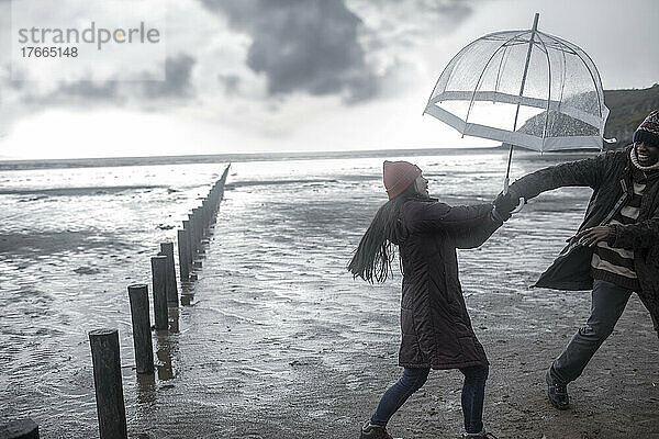 Verspieltes Paar mit Regenschirm am nassen Winterstrand