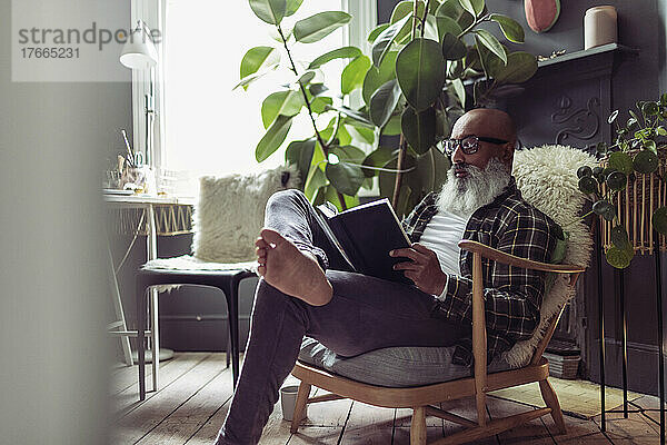 Barfuß reifer Mann mit Bart liest Buch zu Hause