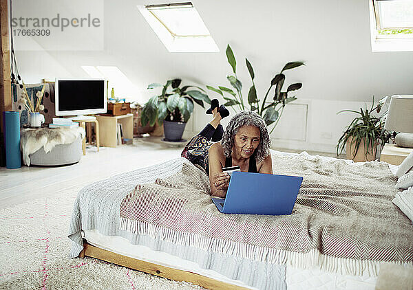 Ältere Frau mit Kreditkarte bezahlt Rechnungen am Laptop auf dem Bett