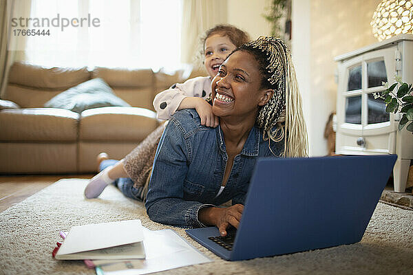Tochter liegt auf glücklicher Mutter  die am Laptop auf dem Boden zu Hause arbeitet