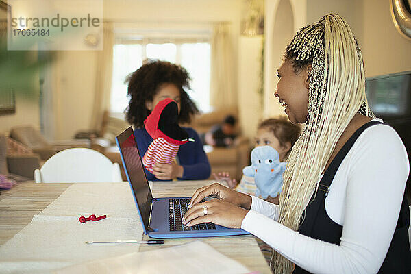 Mutter arbeitet am Laptop und beobachtet Töchter beim Spielen mit Puppen