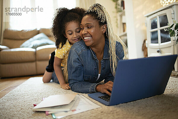 Neugierige Tochter beobachtet Mutter bei der Arbeit am Laptop auf dem Wohnzimmerboden