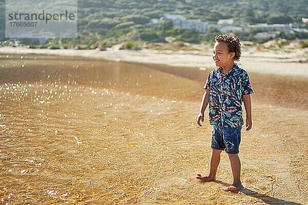 Glücklicher Junge mit Down-Syndrom  der am sonnigen Strand in der Meeresbrandung watet