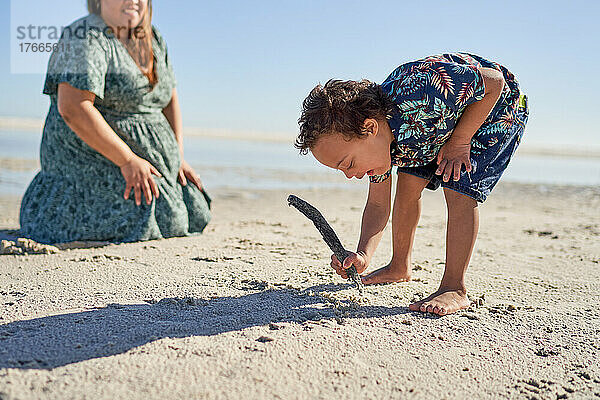 Junge mit Down-Syndrom zeichnet mit Stock im Sand am sonnigen Strand